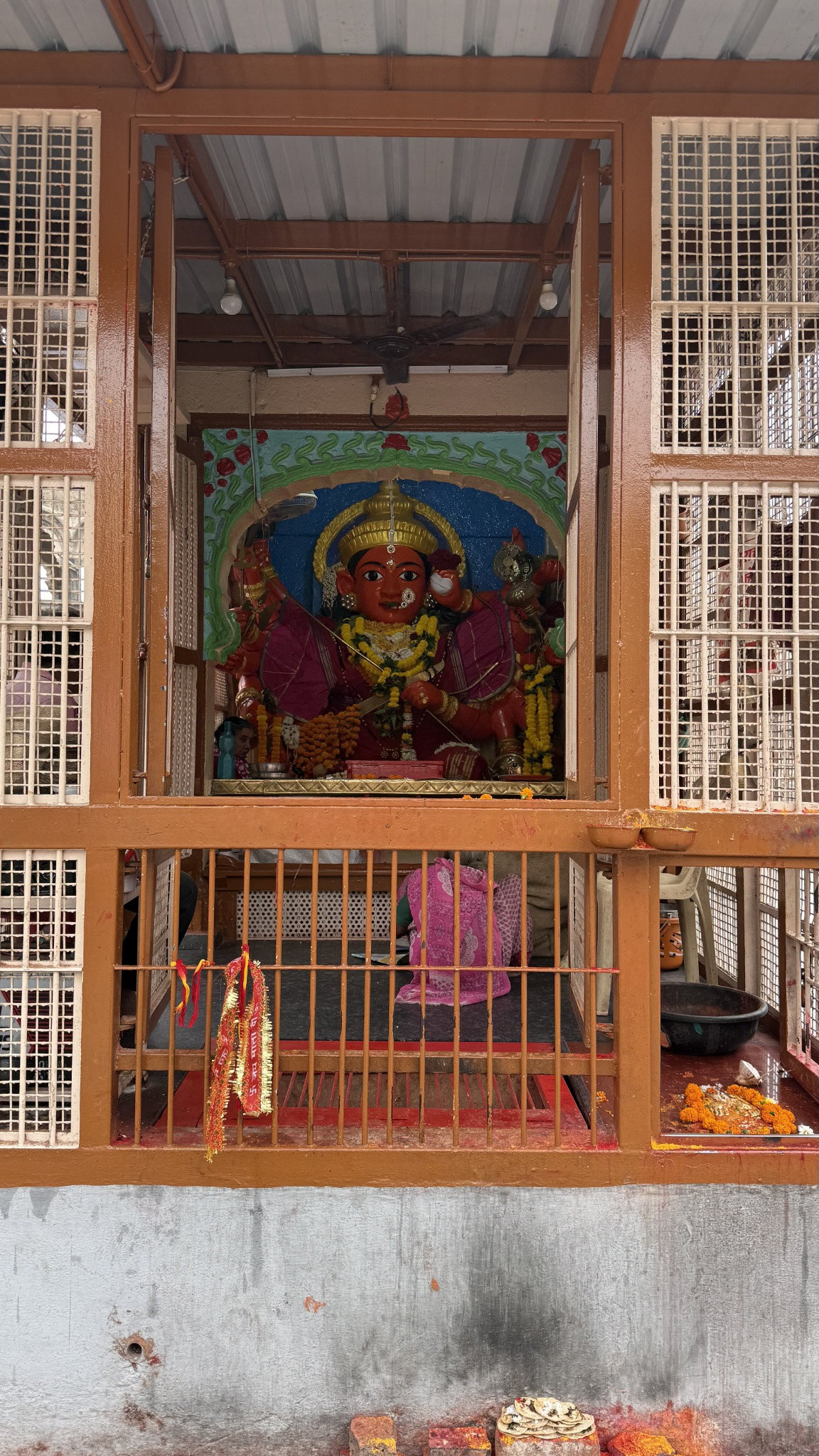 Flower-adorned deity in a Panchavati shrine, framed by a metal-grill doorway.